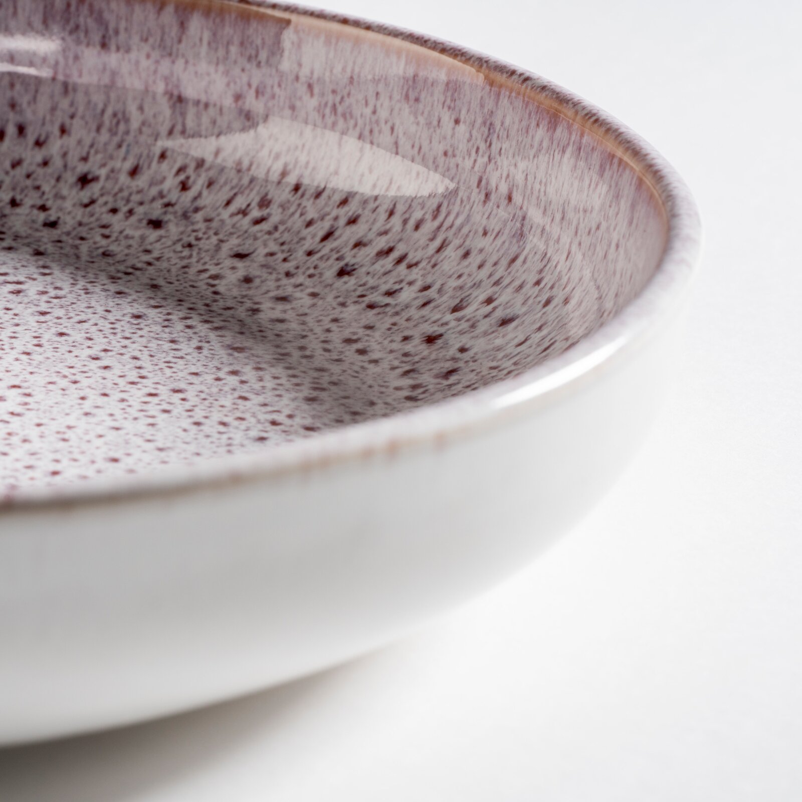 Nahaufnahme einer glasierten Keramikschale mit gesprenkelter Oberfläche in Weiß und Violett, schräg von oben fotografiert (Detailansicht am Rand) auf hellem Hintergrund