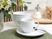 Weiße Porzellantasse auf Untertasse mit Löffel im Vordergrund, dahinter gestapelte weiße Schalen auf einem Tisch; seitliche Perspektive in hellem Küchenambiente