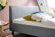 Seitliche Ansicht eines modernen Polsterbetts in Grau mit Kopfteil, dekoriert mit bunten Kissen und weißer Bettwäsche, vor einer rosa Wand.