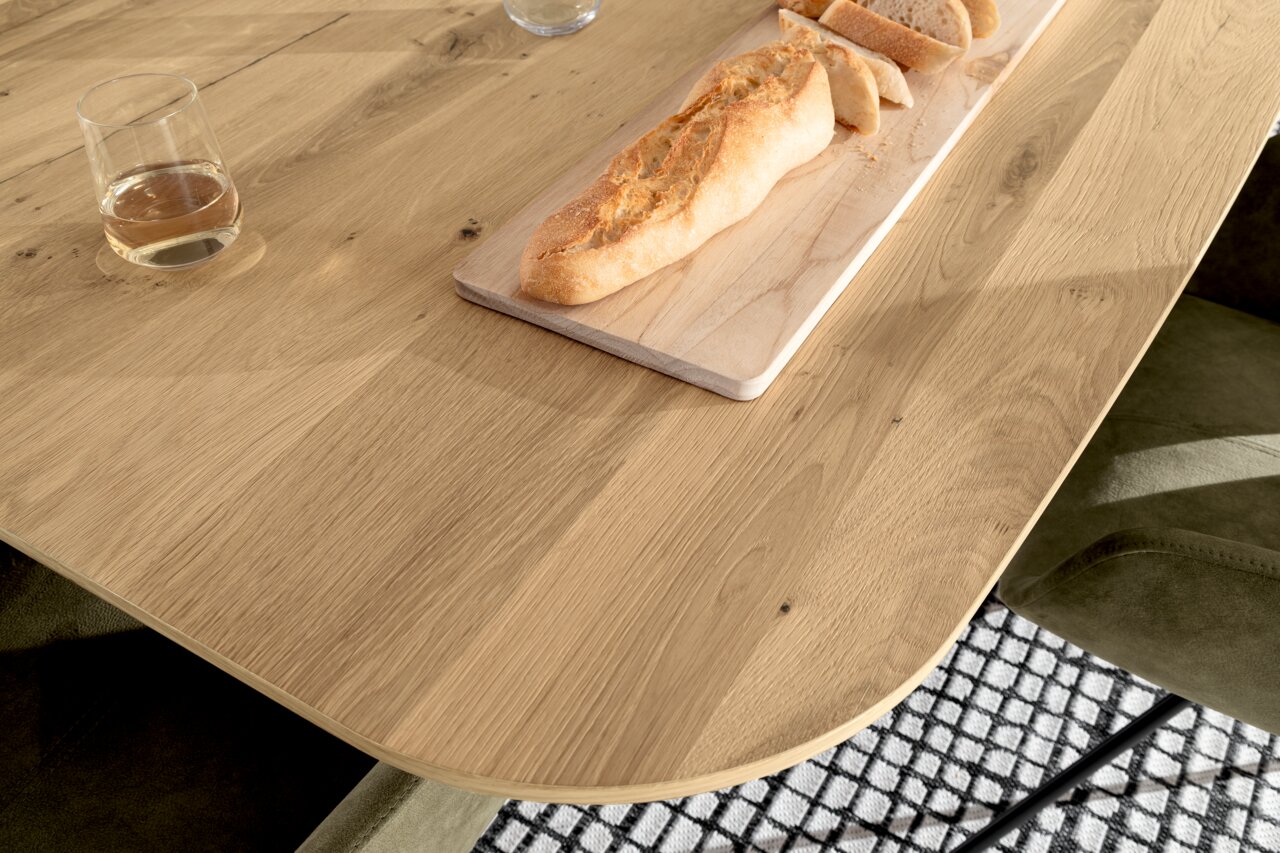 Nahaufnahme eines Venjakob Esstisches ET613 aus Holz mit abgerundeten Ecken, darauf ein Glas und ein Baguette auf einem Schneidebrett. Perspektive von oben.