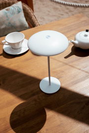 Weiße, minimalistische kabellose LED-Tischleuchte mit rundem Schirm und schlankem Stabfuß auf einem Holztisch neben Tasse und Teekanne, fotografiert aus leicht erhöhter schräger Perspektive.
