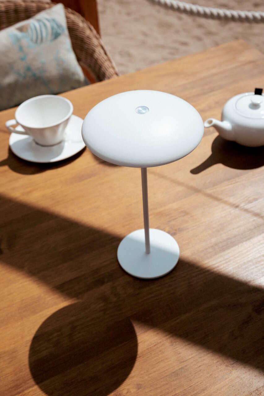 Witte, minimalistische draadloze led-tafellamp met een ronde kap en een slanke staande voet op een houten tafel naast een kopje en een theepot, gefotografeerd vanuit een licht verhoogd, schuin perspectief.