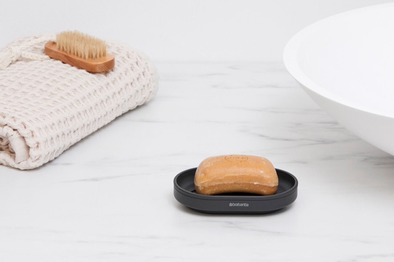 Schwarze Seifenschale mit einem Stück Seife, seitliche Perspektive, auf einem weißen Marmoruntergrund neben einem gefalteten Handtuch und einer Bürste.