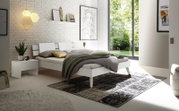 Modernes Schlafzimmer mit einem weißen Bett mit Holz- und Metallkopfteil, seitlicher Blick. Das Zimmer ist hell und minimalistisch eingerichtet mit großen Fenstern, einem Nachttisch und stilvollen Dekorationen.