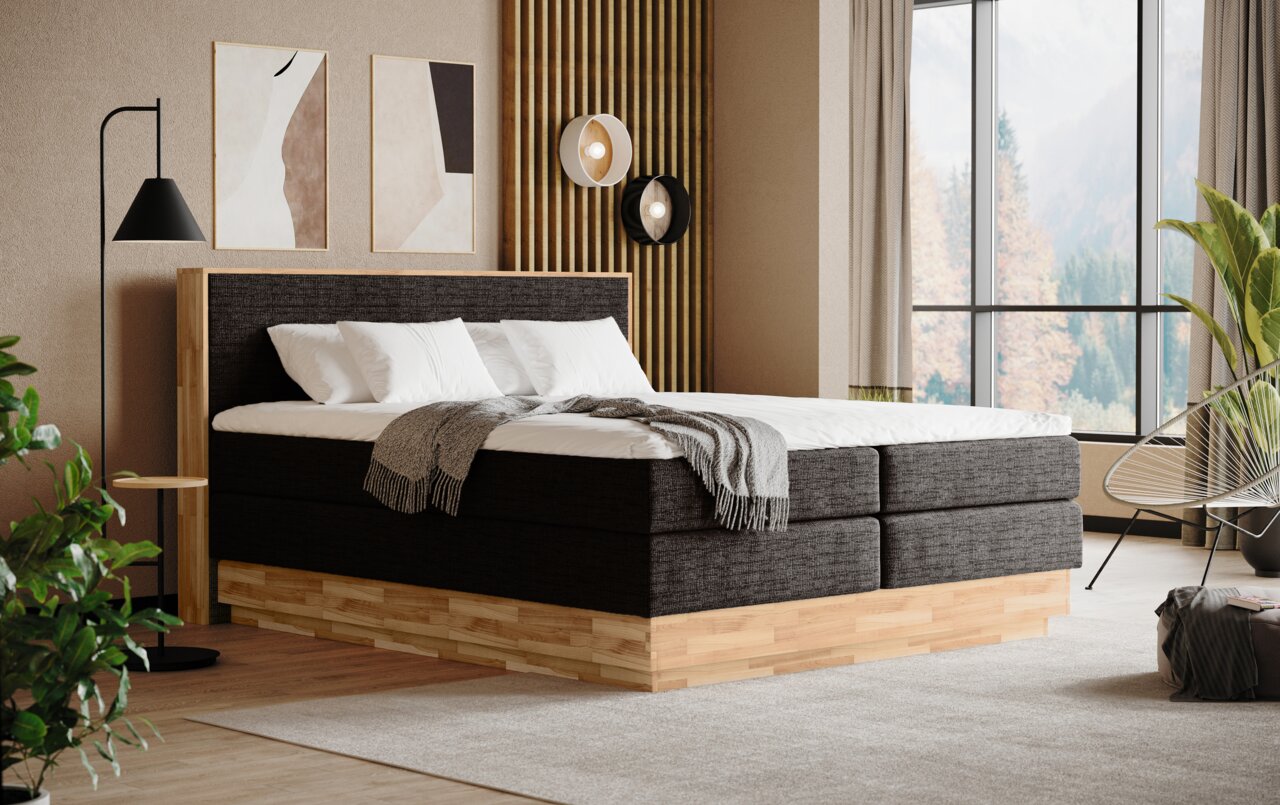 Boxspringbed in een moderne slaapkamer, gezien vanaf de zijkant, met houten frame en grijze stoffen bekleding, versierd met kussens en een deken.