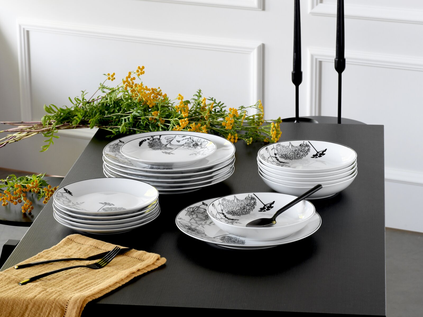 Weißes Porzellan-Geschirrset mit schwarzem floralen Dekor (Teller und Schalen) auf schwarzem Tisch, dekoriert mit gelben Blumen und schwarzem Besteck, Ansicht schräg von vorne leicht von oben.