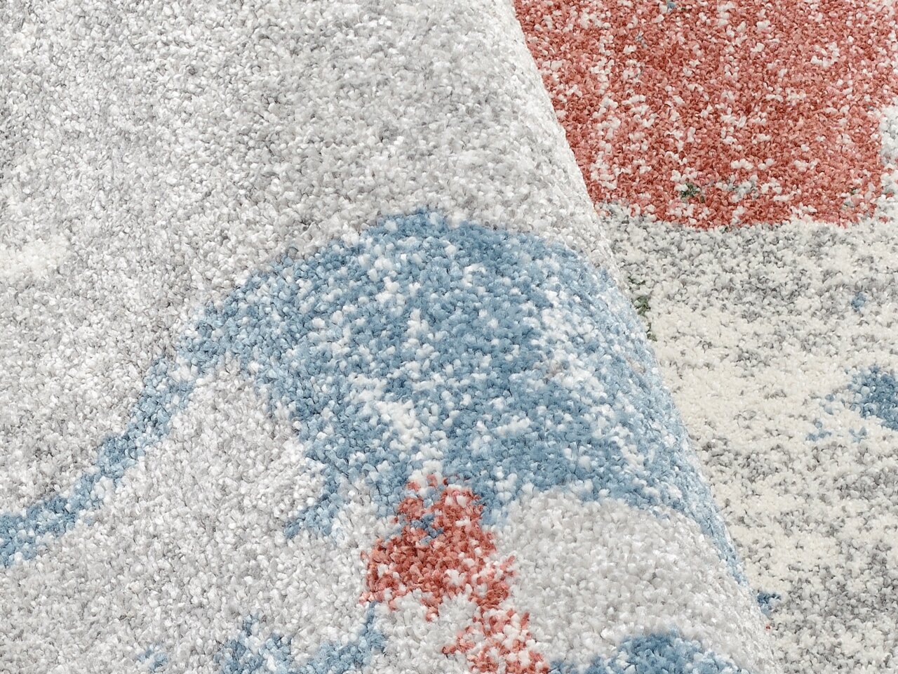 Nahaufnahme eines Frisé-Teppichs mit gemusterten Bereichen in Blau, Rot und Grau. Der Teppich ist teilweise aufgerollt, was die Textur und das Muster aus der Seitenperspektive zeigt.