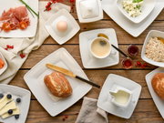 Vogelperspektive auf ein stilvoll gedecktes Frühstückstisch-Arrangement mit weißem Kombiservice, bestehend aus quadratischen Tellern, Tassen und Schalen, gefüllt mit verschiedenen Speisen wie Brot, Käse, Aufschnitt, Müsli und Kaffee.