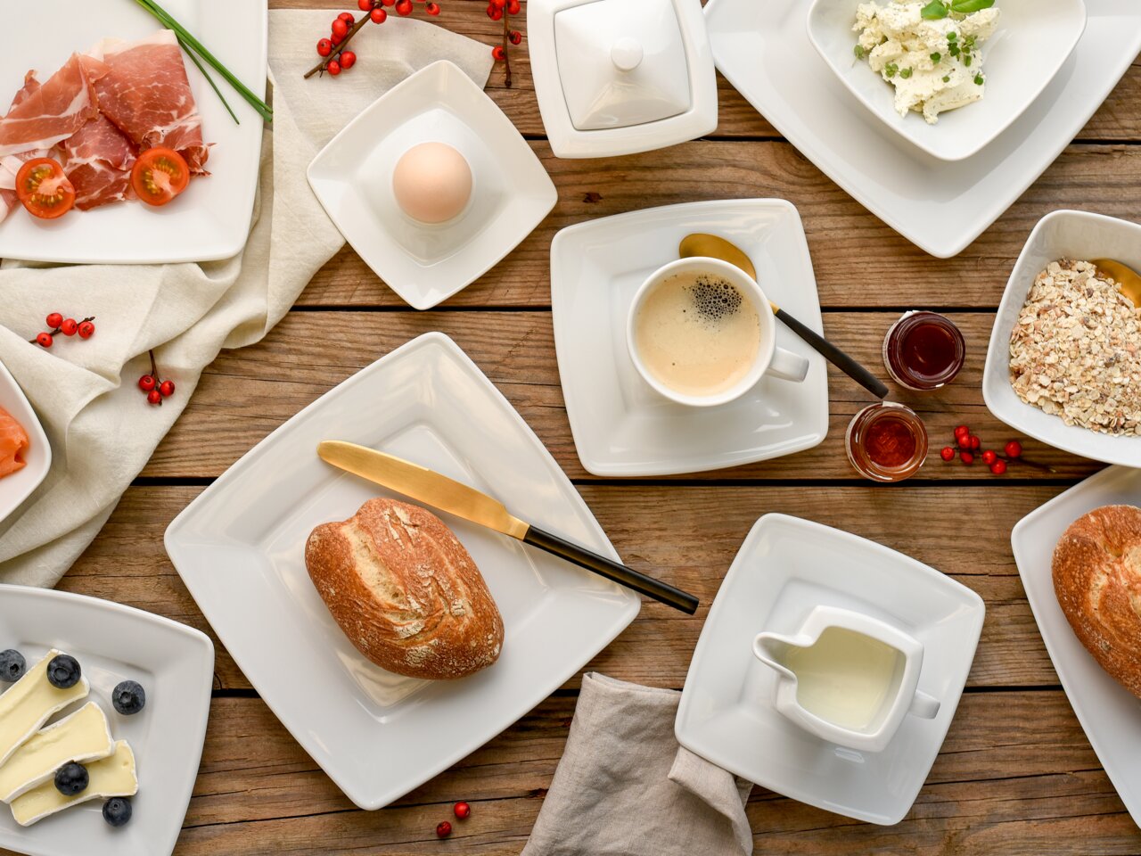 Vogelperspektive auf ein stilvoll gedecktes Frühstückstisch-Arrangement mit weißem Kombiservice, bestehend aus quadratischen Tellern, Tassen und Schalen, gefüllt mit verschiedenen Speisen wie Brot, Käse, Aufschnitt, Müsli und Kaffee.