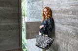 Frau mit rotem Haar lehnt an einer Betonwand und hält eine Shopper-Tasche mit Leopardenmuster in Schwarz-Weiß. Die Tasche wird von vorne gezeigt.
