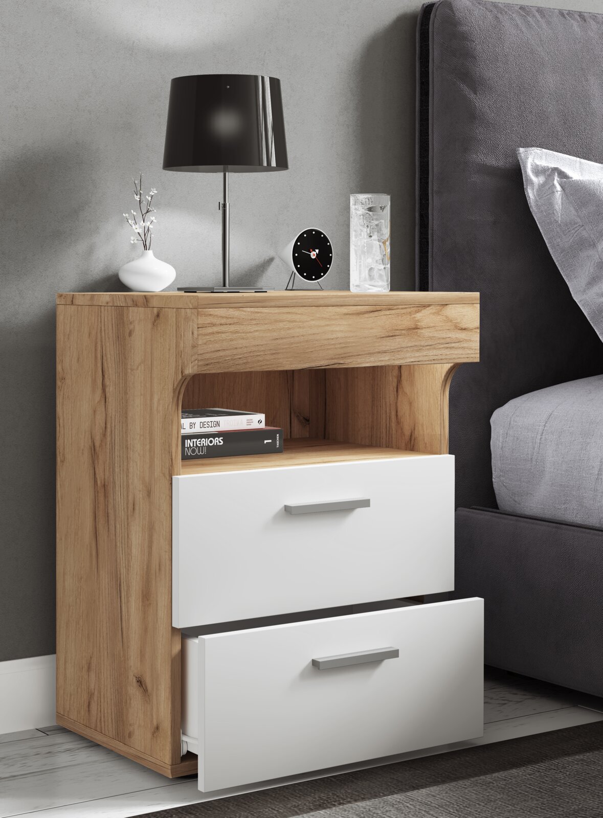 Moderne Nachtkommode in Holzoptik mit zwei weißen Schubladen und offenem Fach, neben einem Bett, fotografiert in schräger Frontansicht auf der Ablage stehen Tischlampe, Uhr, Vase und Glas.