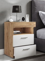 Moderne Nachtkommode in Holzoptik mit zwei weißen Schubladen und offenem Fach, neben einem Bett, fotografiert in schräger Frontansicht; auf der Ablage stehen Tischlampe, Uhr, Vase und Glas.