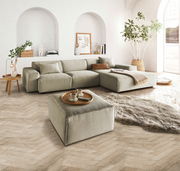 Ein quadratischer, handgefertigter Hocker in einem modernen Wohnzimmer, aus einer leicht erhöhten Perspektive fotografiert. Der Hocker ist in einem neutralen Farbton gehalten und steht auf einem hölzernen Fischgrätboden, umgeben von einem großen Sofa, ein