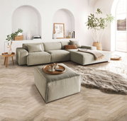 Ein quadratischer, handgefertigter Hocker in einem modernen Wohnzimmer, aus einer leicht erhöhten Perspektive fotografiert. Der Hocker ist in einem neutralen Farbton gehalten und steht auf einem hölzernen Fischgrätboden, umgeben von einem großen Sofa, ein