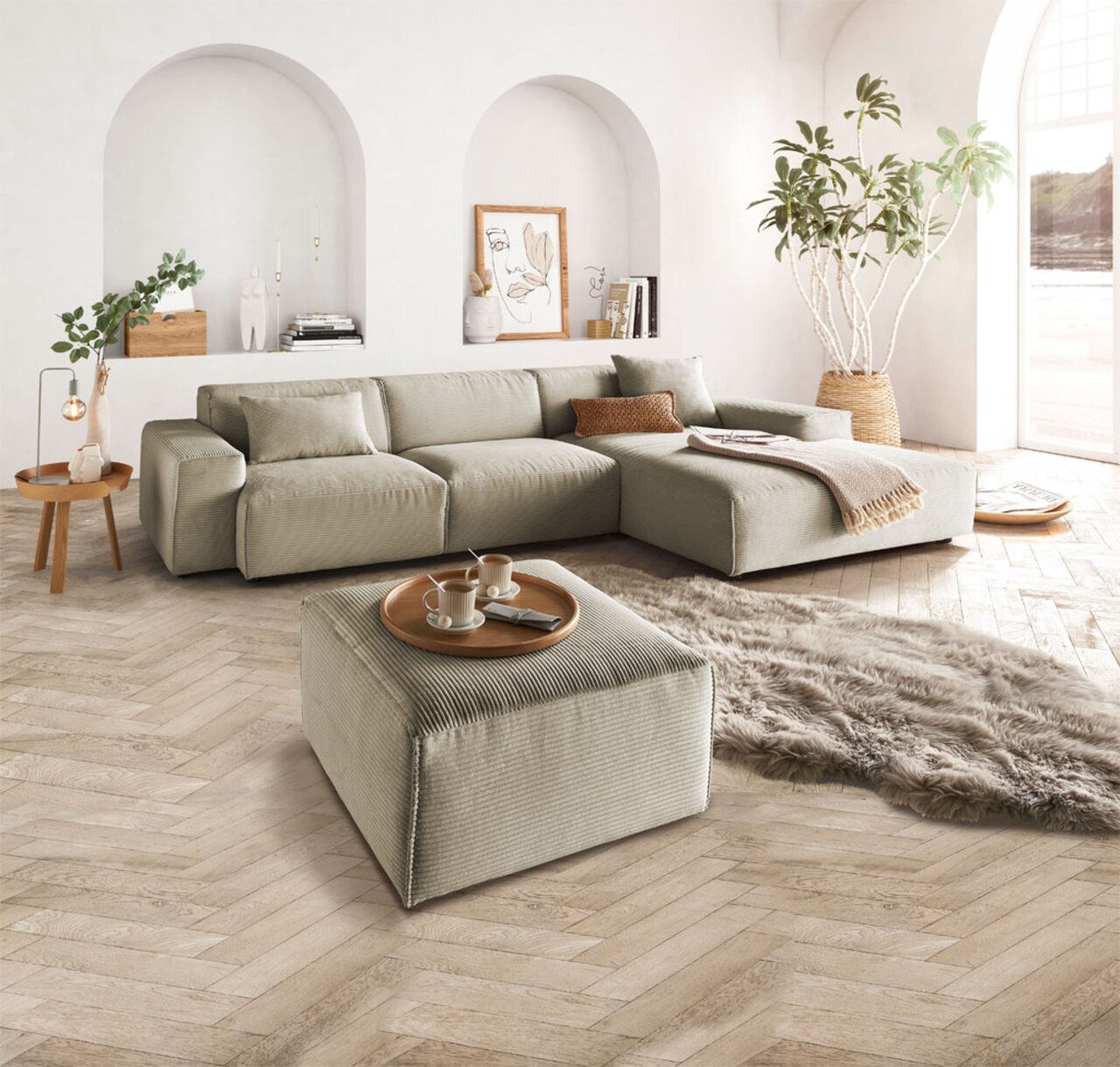 Ein quadratischer, handgefertigter Hocker in einem modernen Wohnzimmer, aus einer leicht erhöhten Perspektive fotografiert. Der Hocker ist in einem neutralen Farbton gehalten und steht auf einem hölzernen Fischgrätboden, umgeben von einem großen Sofa, ein