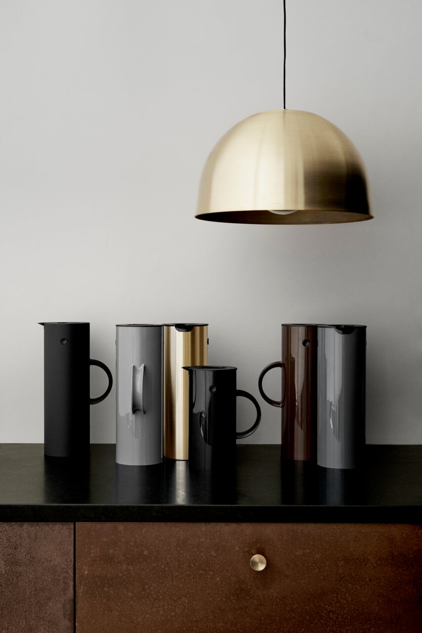 Fünf stilvolle Isolierkannen in verschiedenen Farben, darunter schwarz, grau, gold, braun und dunkelgrau, stehen auf einer dunklen Arbeitsfläche. Eine goldene Hängelampe ist darüber zu sehen. Die Perspektive zeigt die Kannen von vorne.