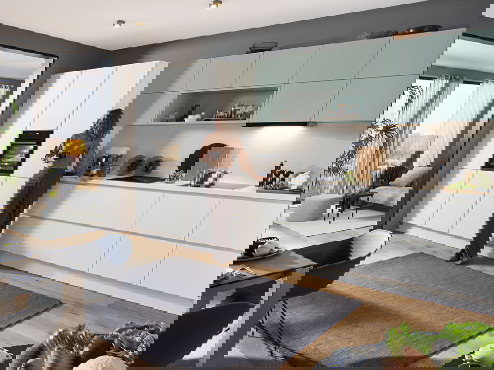 Eine moderne Küche mit mintgrünen Oberschränken und einem großen Fenster. Eine Frau lehnt an einem Hochschrank.