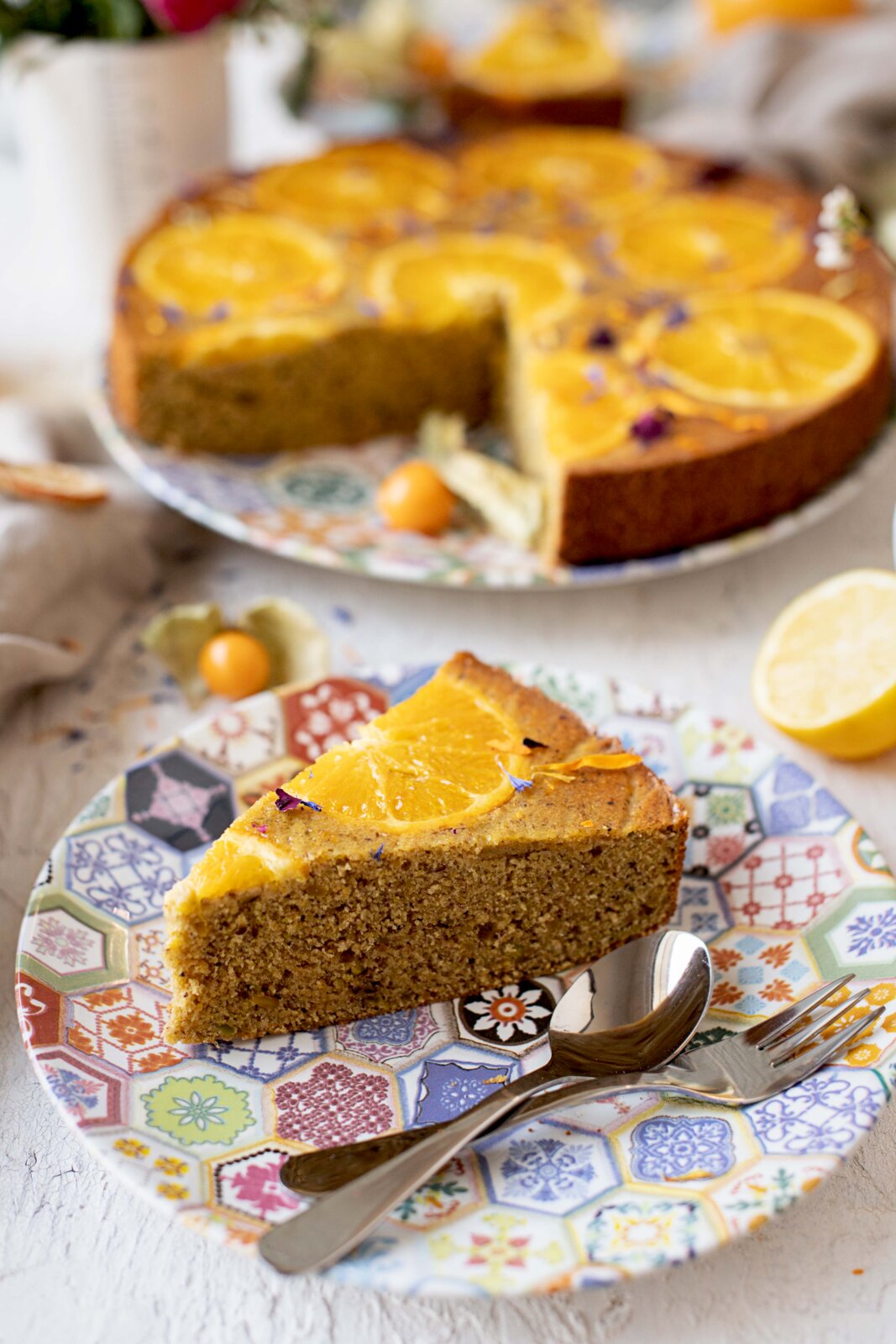 Saftiger Orangenkuchen mit Orangen-Scheiben und Blüten dekoriert, ein Stück auf gemustertem Teller mit Besteck im Vordergrund; Perspektive: leicht von oben mit Fokus auf das Kuchenstück, ganzer Kuchen unscharf im Hintergrund.