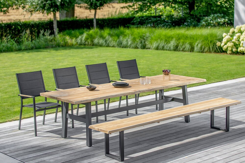 Niehoff-Garden Bank TAVO Moderner Esstisch und Bank aus Holz mit Metallgestell, umgeben von schwarzen Stühlen, auf einer Terrasse mit Blick auf einen grünen Garten, seitliche Perspektive.