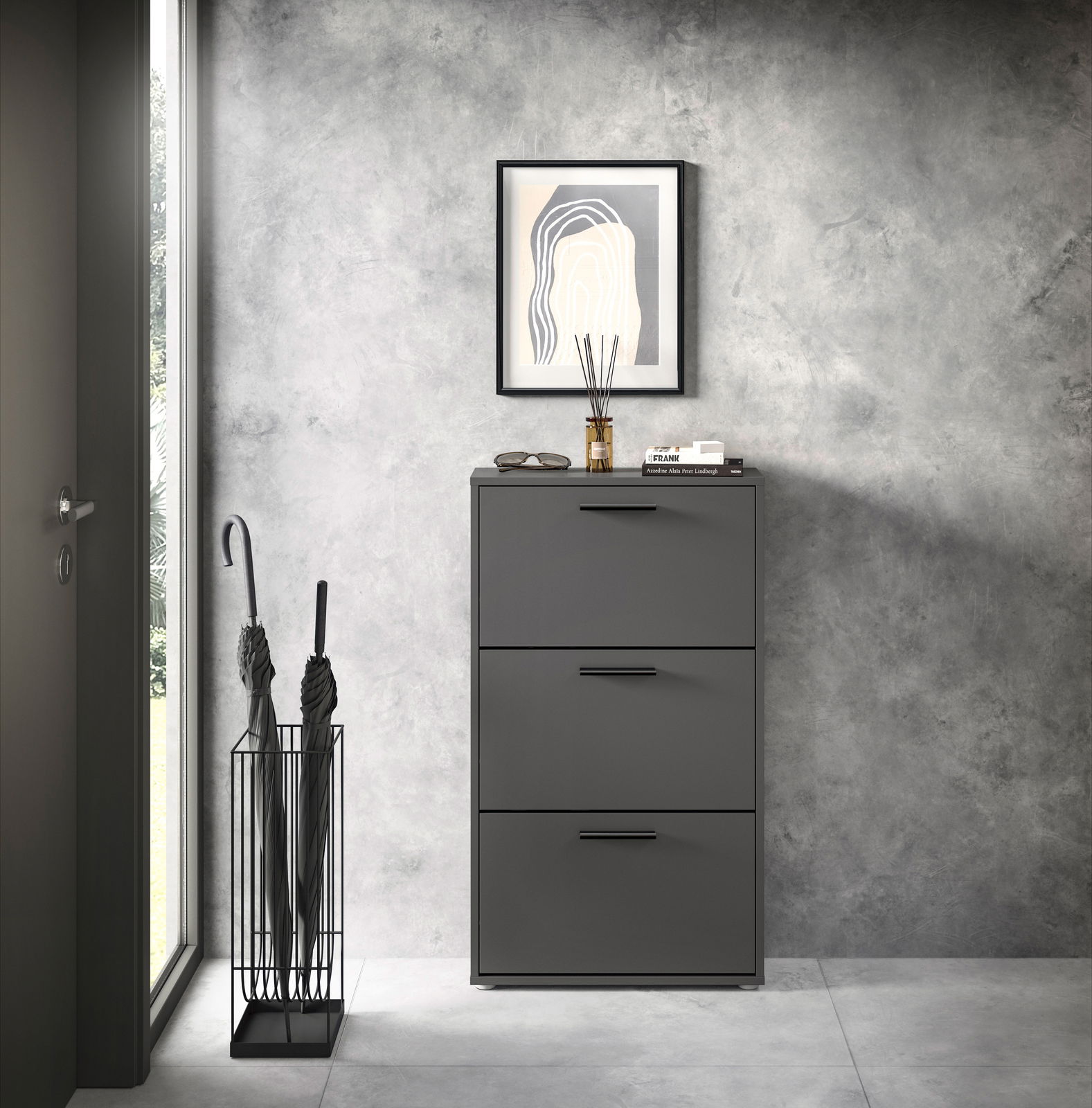 Moderner Schuhkipper mit drei Klappen in dunklem Grau, frontal fotografiert, neben einem Regenschirmständer und unter einem gerahmten Bild an einer Betonwand.
