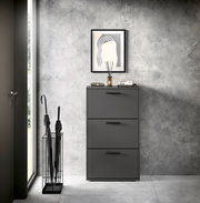 Moderner Schuhkipper mit drei Klappen in dunklem Grau, frontal fotografiert, neben einem Regenschirmständer und unter einem gerahmten Bild an einer Betonwand.