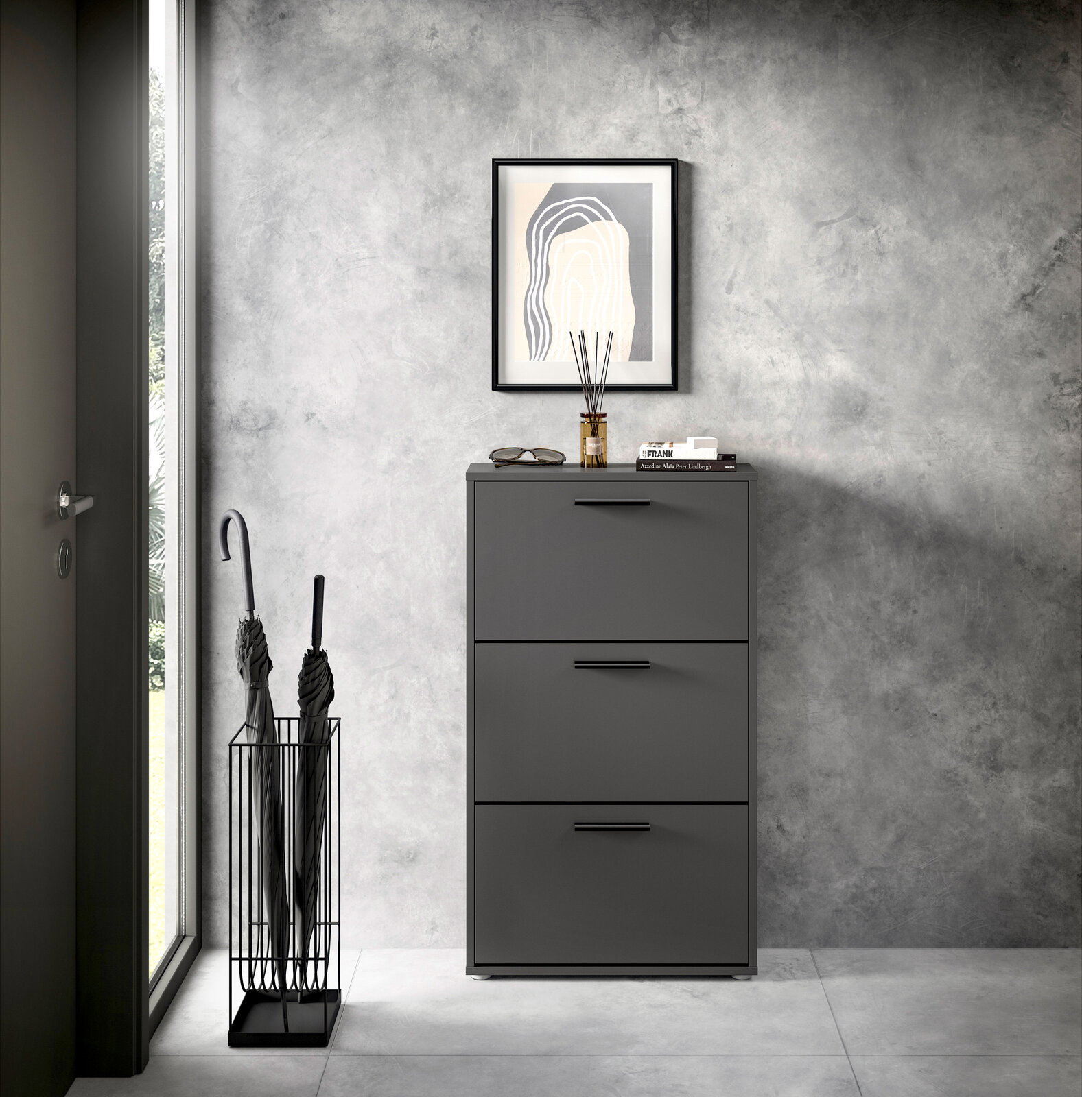 Moderner Schuhkipper mit drei Klappen in dunklem Grau, frontal fotografiert, neben einem Regenschirmständer und unter einem gerahmten Bild an einer Betonwand.