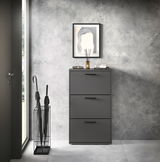 Moderner Schuhkipper mit drei Klappen in dunklem Grau, frontal fotografiert, neben einem Regenschirmständer und unter einem gerahmten Bild an einer Betonwand.