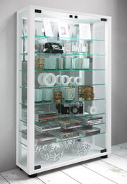 Weiße Glasvitrine mit Glasböden und transparenten Schiebetüren, gefüllt mit Dekoartikeln, CDs und Kamera; Perspektive: frontale Ansicht mit leichtem Blick von links oben.