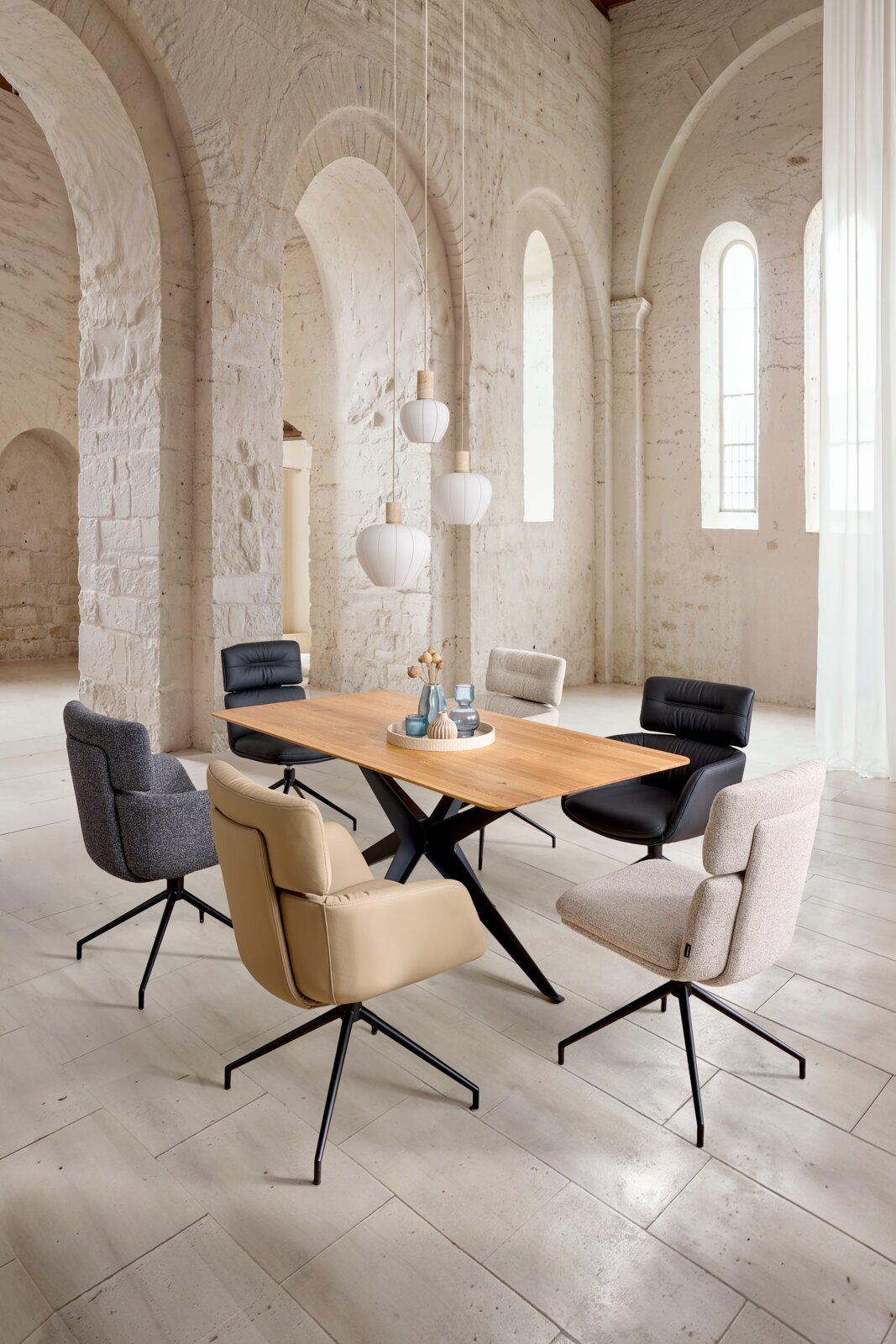 Schräg von vorne fotografierter moderner Esstisch aus Holz mit schwarzem Kreuzgestell, umgeben von gepolsterten Drehstühlen in Beige, Grau und Schwarz in einem hellen Raum mit hohen Rundbogenfenstern und drei weißen Pendelleuchten.