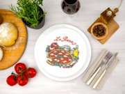 Draufsicht auf einen weißen Teller mit „La Pizza“-Motiv und bunter Pizza-Illustration, arrangiert auf hellem Tisch mit Tomaten, Teig, Kräutertopf, Rotweinglas, Öl, Pfeffer und Besteck.