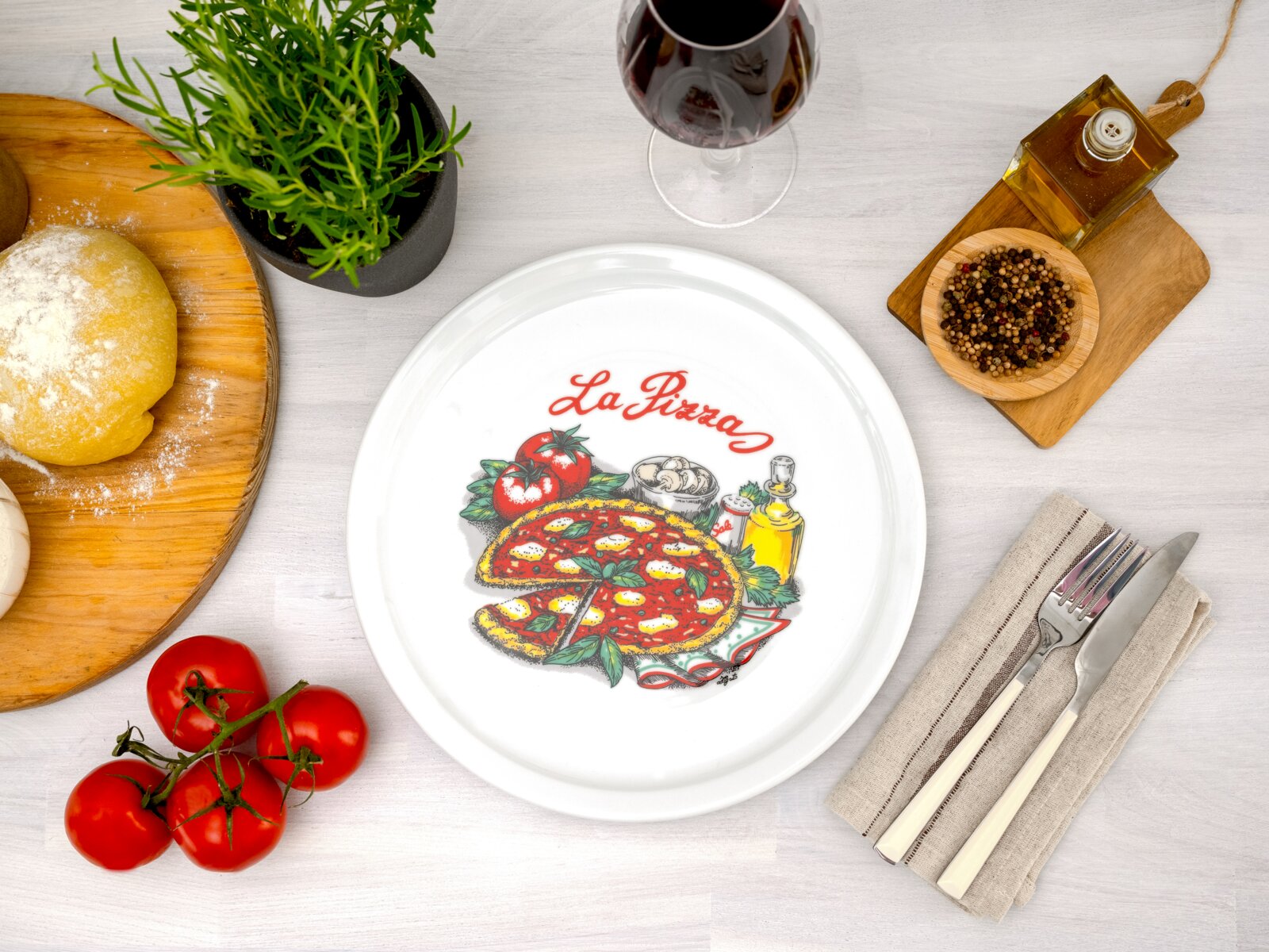 Draufsicht auf einen weißen Teller mit „La Pizza“-Motiv und bunter Pizza-Illustration, arrangiert auf hellem Tisch mit Tomaten, Teig, Kräutertopf, Rotweinglas, Öl, Pfeffer und Besteck.