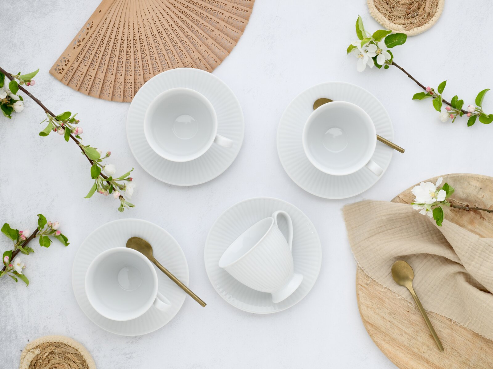 Draufsicht auf vier weiße Porzellan-Kaffeetassen mit Untertassen, teils mit goldfarbenen Löffeln, arrangiert auf hellem Hintergrund mit Blütenzweigen, Holzbrett, Stoffserviette und dekorativem Fächer.