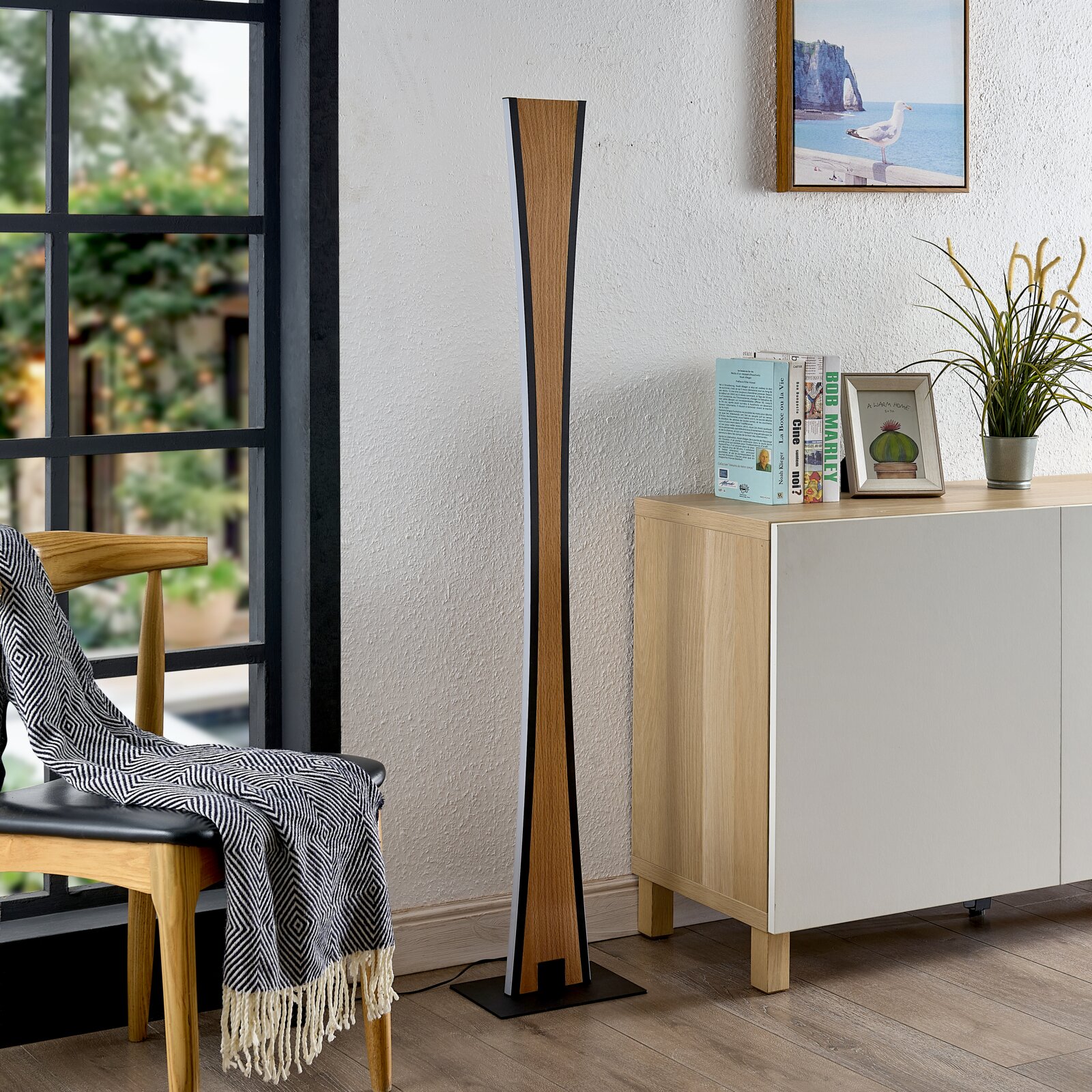 Hoher, schlanker Standlautsprecher in Holzoptik mit schwarzen Seiten und quadratischem Standfuß neben einem Sideboard im Wohnzimmer, frontal aus leichter Seitenperspektive fotografiert