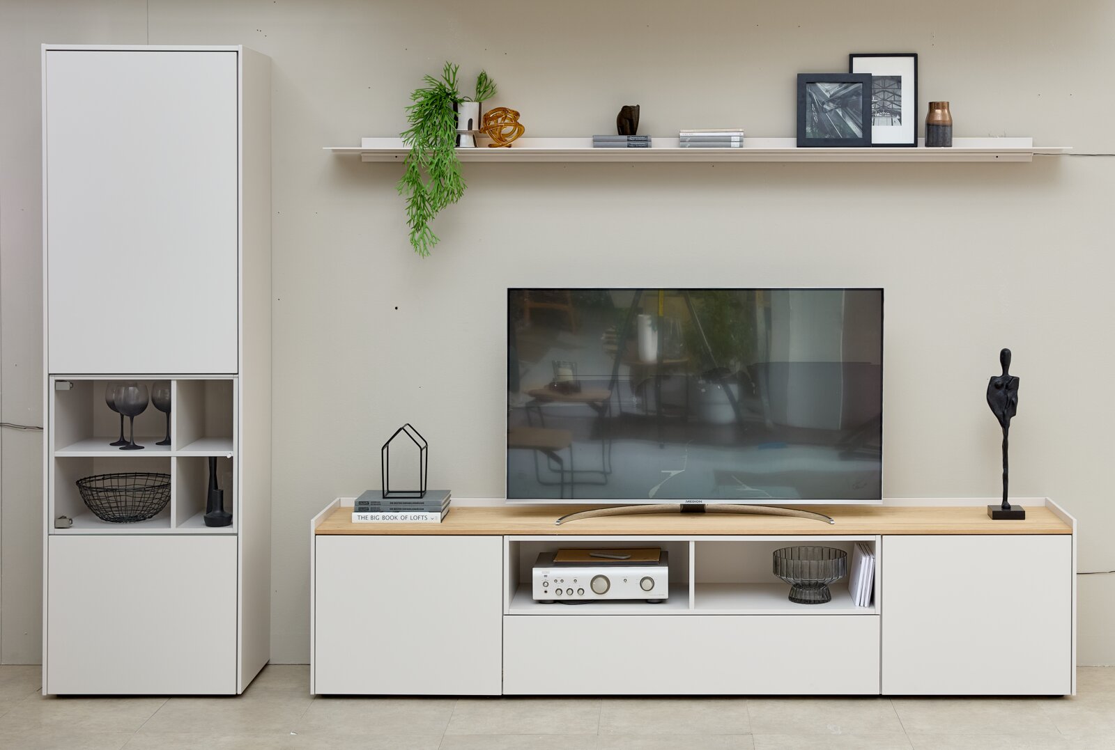 Frontaal aanzicht van een moderne witte tv-wandcombinatie met hoge vitrinekast, laag tv-meubel met houtkleurig blad en open vakken, en een lange wandplank met decoratie.