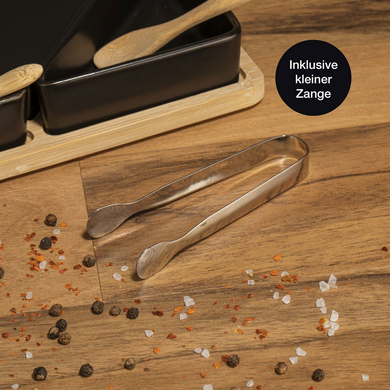 Nahaufnahme eines Serviersets aus Bambus mit fünf schwarzen Schalen, kleinen Holzlöffeln und einer kleinen Zange auf einem Holztisch, umgeben von Gewürzen.