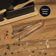 Nahaufnahme eines Serviersets aus Bambus mit fünf schwarzen Schalen, kleinen Holzlöffeln und einer kleinen Zange auf einem Holztisch, umgeben von Gewürzen.