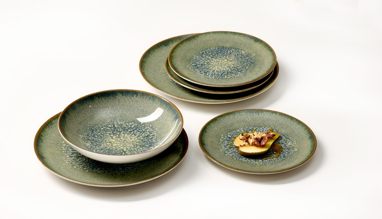 Groenblauw keramisch serviesset met organisch gespikkeld glazuur en bruine randen, schuin van boven gefotografeerd bestaande uit gestapelde borden en een kom, met een klein gerecht op één bord.