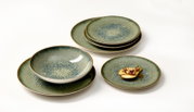 Groenblauw keramisch serviesset met organisch gespikkeld glazuur en bruine randen, schuin van boven gefotografeerd; bestaande uit gestapelde borden en een kom, met een klein gerecht op één bord.