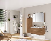 Moderner Badezimmer-Mittelschrank in Holzoptik, links an der Wand montiert, mit dekorativen Elementen darauf, aus seitlicher Perspektive fotografiert.