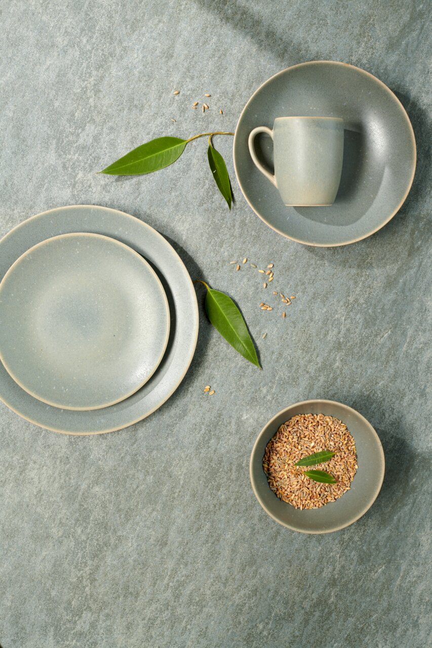 Vogelperspektive auf ein elegantes, graues Kombiservice-Set mit Teller, Tasse und Schale, dekoriert mit grünen Blättern und Getreidekörnern.
