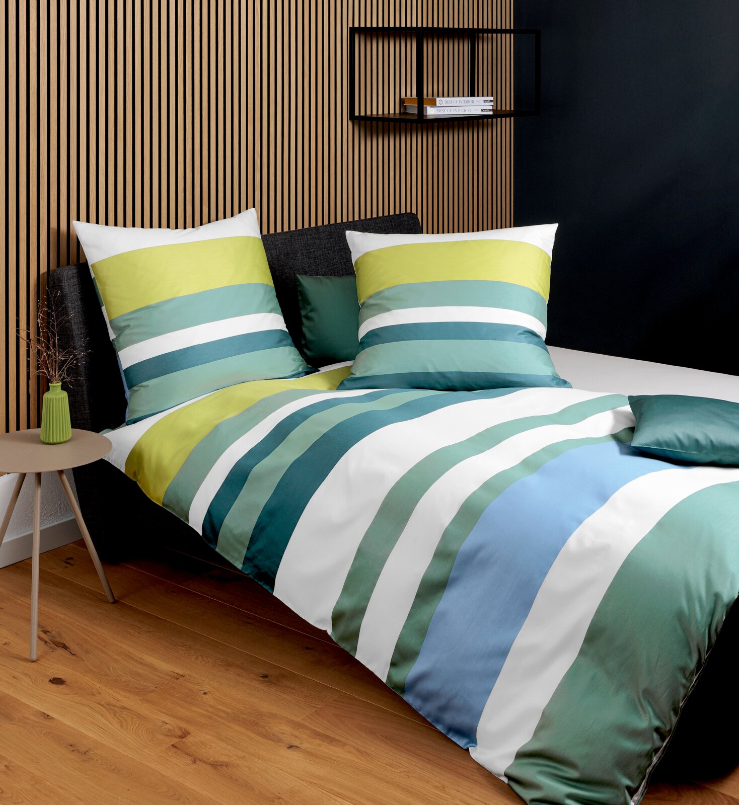 Gestreifte Bettwäsche in Grün-, Blau- und Weißtönen auf einem Bett mit dunklem Kopfteil, fotografiert in schräger Vorderansicht im modern eingerichteten Schlafzimmer