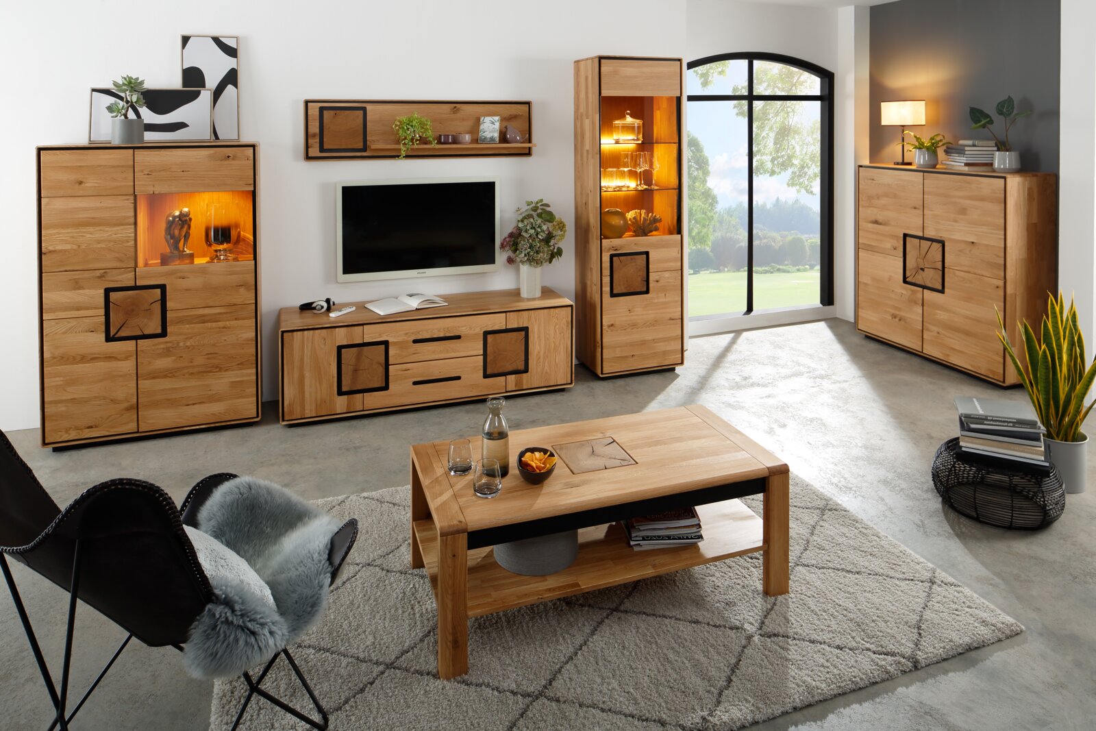 Modernes Wohnzimmer mit Wohnwand und Couchtisch aus hellem Massivholz, inklusive Highboard und Vitrine mit Beleuchtung; Frontansicht in leichter Schrägperspektive von links.
