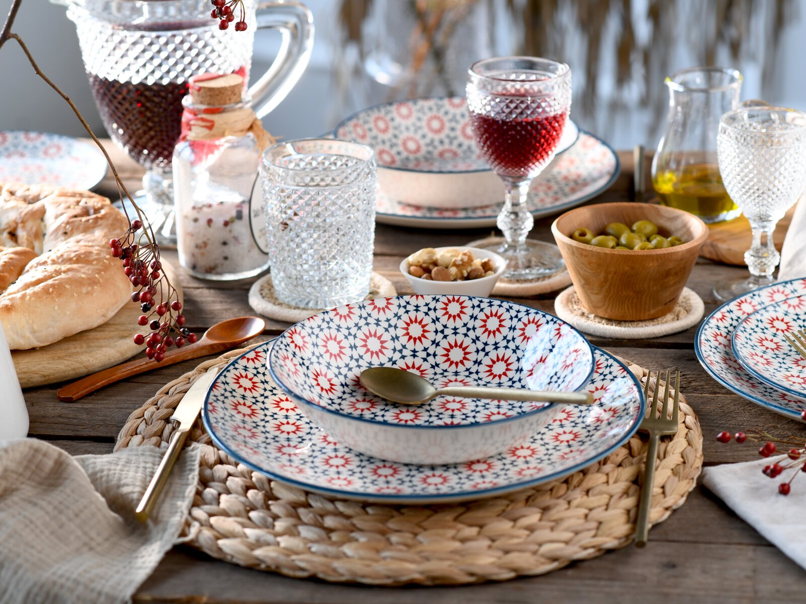 Schräg von oben fotografiertes, gedecktes Tisch-Setting mit gemustertem Keramikgeschirr in Rot-Blau, strukturierten Wassergläsern, Karaffe mit rotem Getränk, Brot, Oliven, Nüssen und Öl auf rustikalem Holztisch.