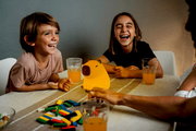 Zwei lachende Kinder sitzen an einem Tisch mit einem leuchtenden Akku-Nachtlicht in Form eines Schweins in der Mitte, umgeben von Getränken und bunten Spielsteinen. Perspektive von vorne.