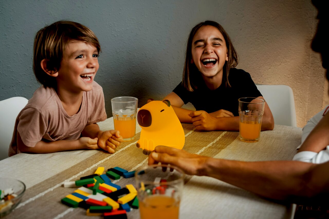 Zwei lachende Kinder sitzen an einem Tisch mit einem leuchtenden Akku-Nachtlicht in Form eines Schweins in der Mitte, umgeben von Getränken und bunten Spielsteinen. Perspektive von vorne.