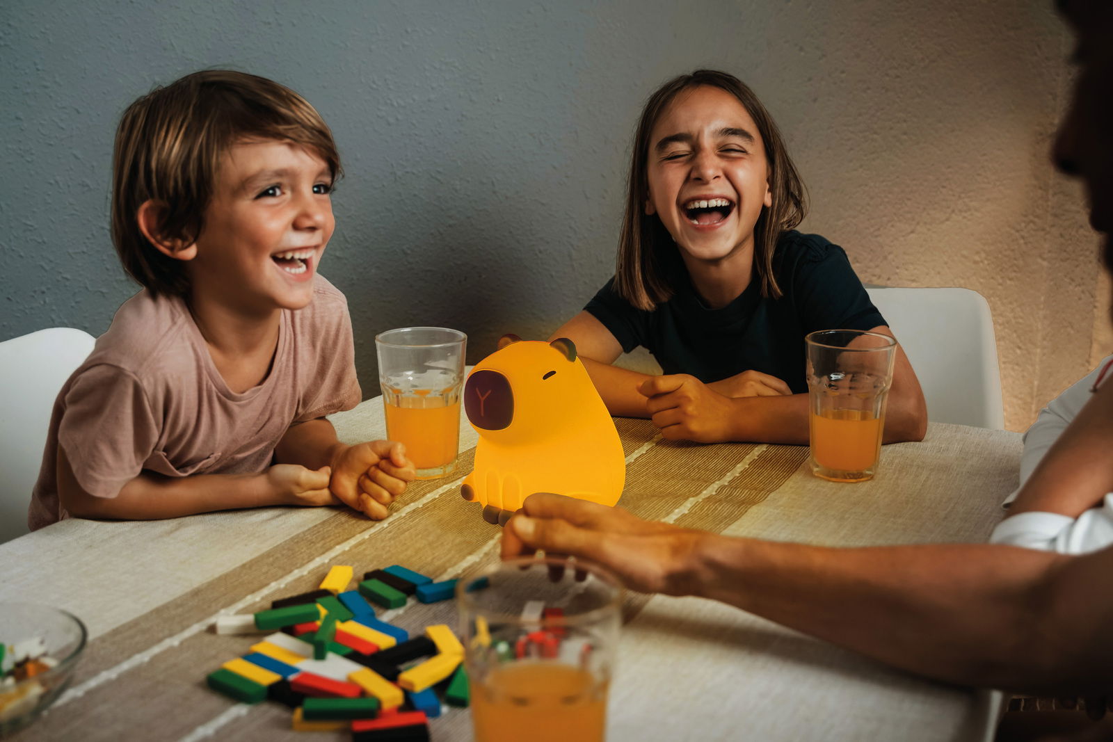 Zwei lachende Kinder sitzen an einem Tisch mit einem leuchtenden Akku-Nachtlicht in Form eines Schweins in der Mitte, umgeben von Getränken und bunten Spielsteinen. Perspektive von vorne.