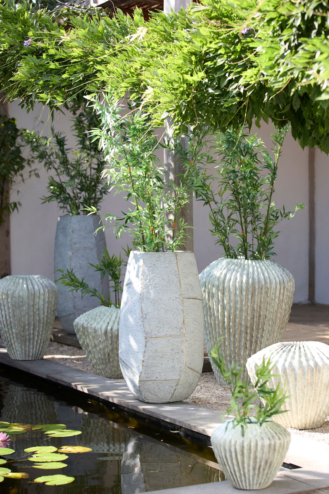 Light&Living Topf AGELO Großer, moderner Pflanztopf aus hellem Material, umgeben von grünen Pflanzen, in einem Garten mit Teich. Perspektive von vorne.