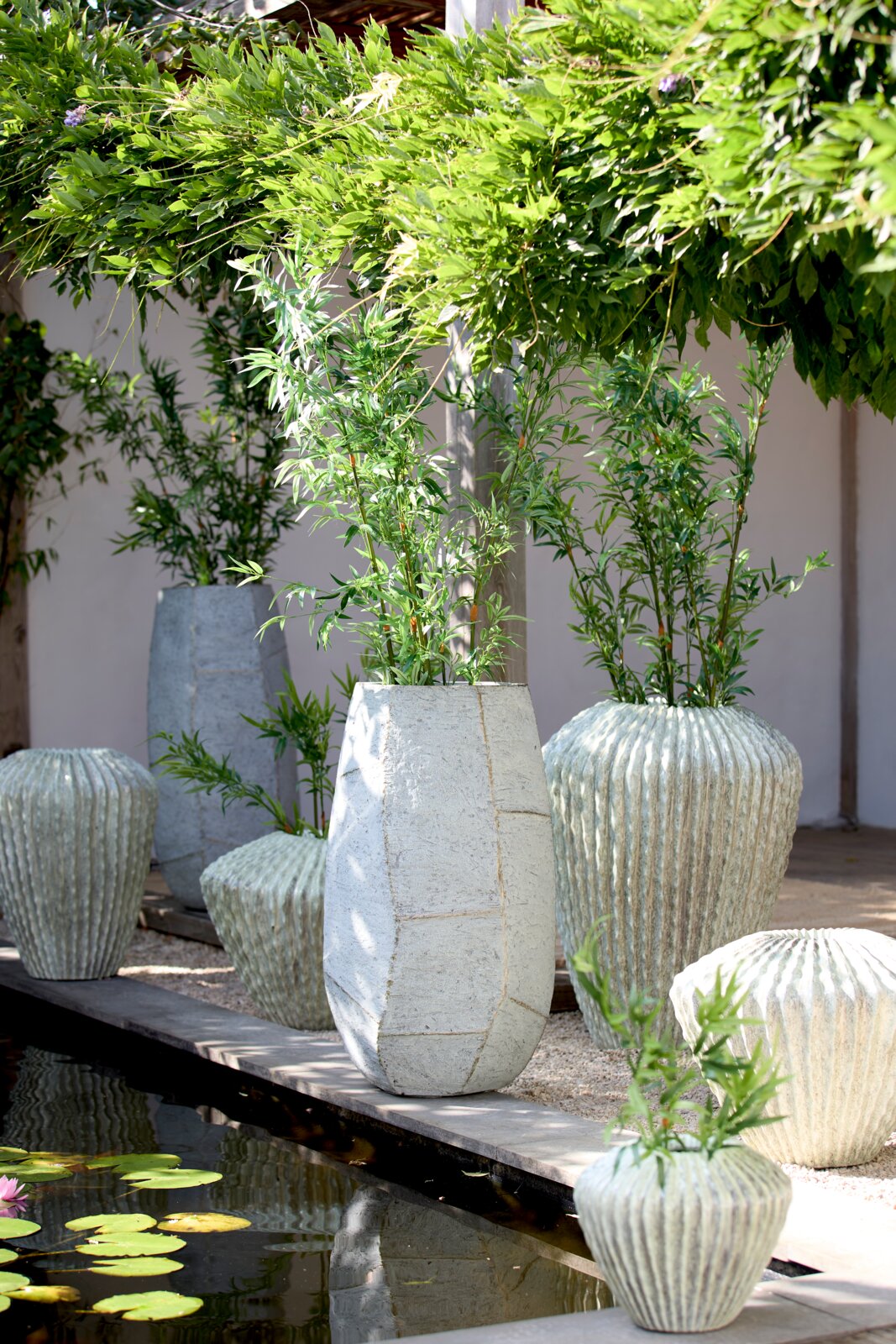Light&Living Pan AGELO 105 cm Grote, moderne plantenpot van licht materiaal, omringd door groene planten, in een tuin met een vijver. Aanzicht van voren.