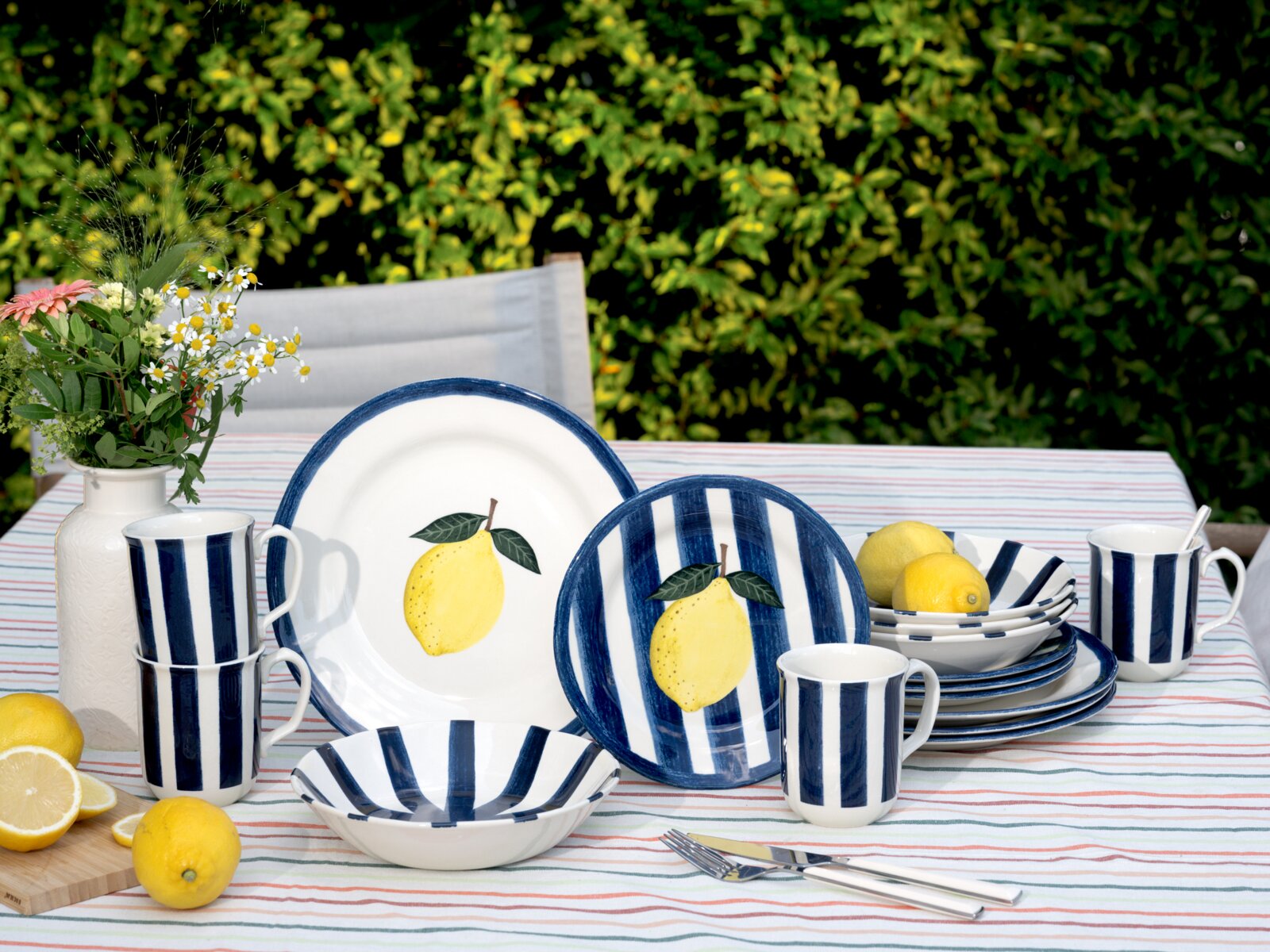 creaTable Kombiservice 16-tlg SUMMER STRIPES Outdoor-Tischgedeck mit blau-weiß gestreiftem Keramikgeschirr und Zitronenmotiv (Teller, Schalen, Becher), Zitronen und Blumenvase auf gestreifter Tischdecke, frontal aus leicht erhöhter Perspektive aufgenommen
