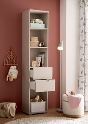 Hohes weißes Kinderzimmer-Regal mit offenen Fächern und zwei herausgezogenen Schubladen, gefüllt mit Büchern, Spielzeug und Aufbewahrungsboxen; seitliche Frontansicht im Raum neben Stehlampe und Sitzpouf.